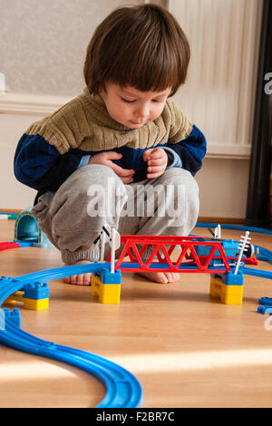 Caucasien enfant, garçon, 3 - 5 ans, à la maison dans la chambre avec parquet au sol, accroupi à Tommy à moteur Réservoir plastique jouets et chemin de fer pont. Banque D'Images