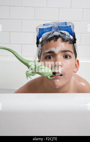 Jeune garçon dans une baignoire avec des lunettes sur le dessus de la tête et dans la bouche dinosaure en plastique avec de l'eau savonneuse examine l'appareil photo
