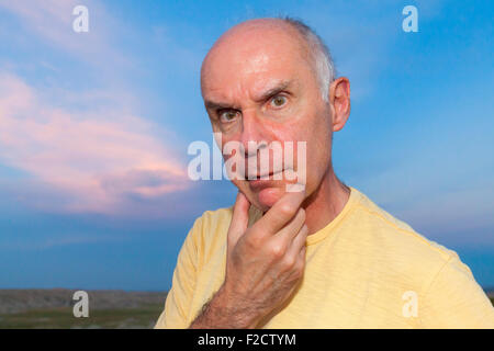 A senior man posing with thoughtful, sérieux et expressions idiotes à l'extérieur contre un ciel bleu. Banque D'Images