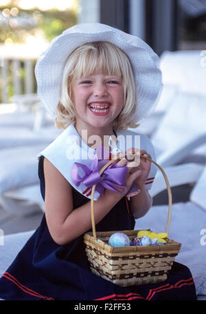 Une jeune blonde américaine pose joyeusement dans sa robe et chapeau de Pâques Pâques avec un panier d'œufs de Pâques colorés et bonbons sur ses genoux. Dressing up et la coloration des oeufs pour fêter les vacances de Pâques, ont longtemps été des traditions pour les enfants aux États-Unis. Banque D'Images