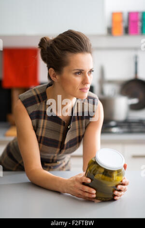 Une femme élégante se penche sur son comptoir de la cuisine, tenant un pot de cornichons, et à la recherche au loin dans la distance. Elle est en pleine réflexion, imaginer toutes les façons qu'elle pourra utiliser les pickles bientôt. Banque D'Images