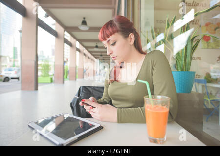 Young handsome young redhead woman using smartphone assis dans un bar, avec un jus et une tablette sur la table, à la baisse, en tapotant sur l'écran - technologie, réseau social, concept multitâche Banque D'Images