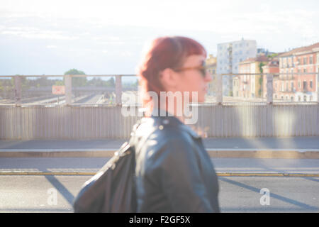 Profil de demi-longueur d'une jeune femme rousse caucasienne beau marcher dans la ville, donnant sur la gauche, pensive - Gravité - concept - rétroéclairage portant une veste en cuir - se concentrer sur l'arrière-plan Banque D'Images