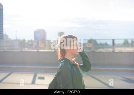 Profil de demi-longueur d'une jeune femme rousse caucasienne beau marcher dans la ville, donnant sur la gauche, pensive - Gravité - concept - rétroéclairage portant une veste en cuir Banque D'Images