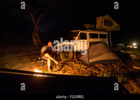 Homme assis par un incendie au téléphone avec son Land Rover dans l'arrière-plan au Botswana, l'Afrique Banque D'Images