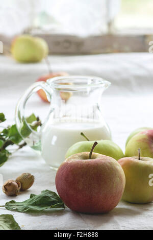 Tas de pommes avec des feuilles, de noisettes et de pot de lait sur nappe blanche avec fenêtre en arrière-plan Banque D'Images