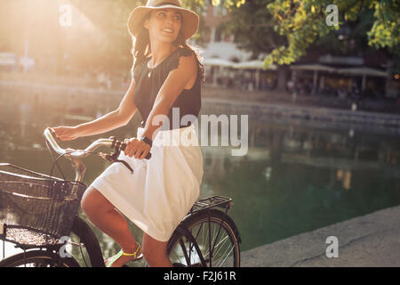 Portrait de belle jeune femme de la bicyclette le long d'une rue près de l'étang sur une journée d'été. Femme portant hat looking away whi Banque D'Images