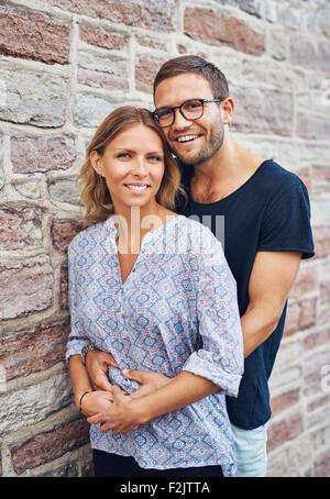 La moitié d'un projectile de corps Sweet Man Hugging son amie de son derrière tout en se penchant contre mur en béton et lui sourit à l'appareil photo Banque D'Images