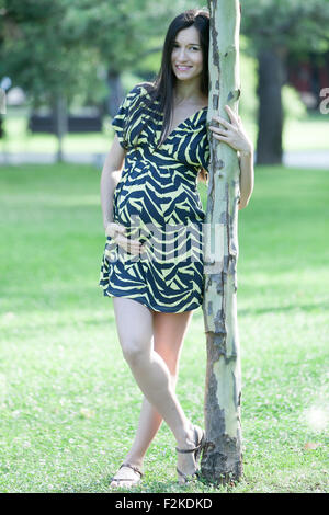 Young pregnant woman smiling et se penchant vers un petit arbre dans le parc. Banque D'Images