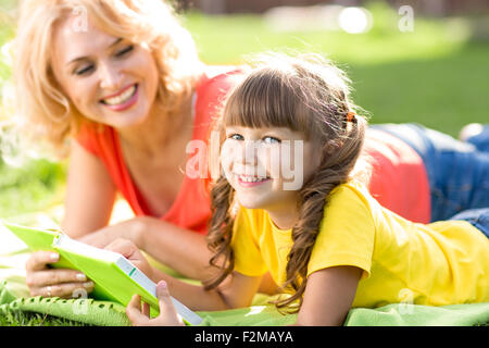 Cute little girl reading book avec sa mère et le sourire. L'herbe d'été en arrière-plan. Banque D'Images