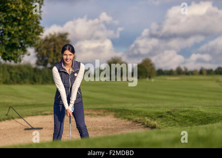 Jeune femme golfeur debout dans un bunker de sable sur un terrain de golf avec un club dans sa main en souriant à la caméra en tant qu'elle tente de quitter Banque D'Images