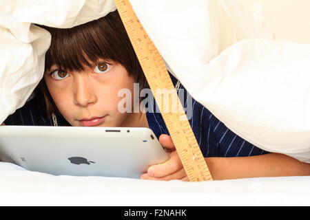 Caucasien enfant, garçon, 10 - 11 ans. La moitié de se cacher sous une couette blanche soutenu par une règle. Tête et épaules de garçon qu'il utilise l'ipad, le contact avec les yeux. Banque D'Images