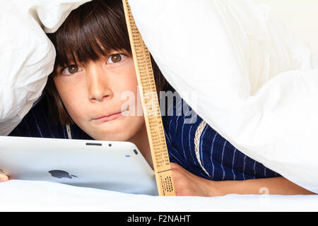 Caucasien enfant, garçon, 10 - 11 ans. La moitié de se cacher sous une couette blanche soutenu par une règle. Tête et épaules de garçon qu'il utilise l'ipad, pensive. Banque D'Images