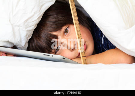 Caucasien enfant, garçon, 10 - 11 ans. La moitié de se cacher sous une couette blanche soutenu par une règle. Tête et épaules de garçon qu'il utilise l'ipad, le contact avec les yeux. Banque D'Images