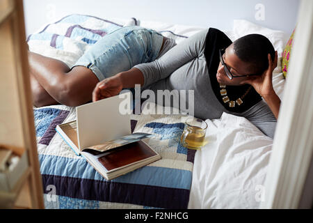 Mixed Race woman boire du thé et de la lecture on bed Banque D'Images