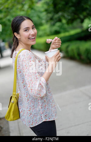 Hispanic woman eating le yogourt glacé sur les trottoirs de la ville Banque D'Images