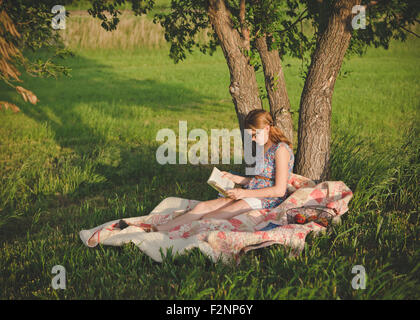 Caucasian girl reading book sous tree in field Banque D'Images