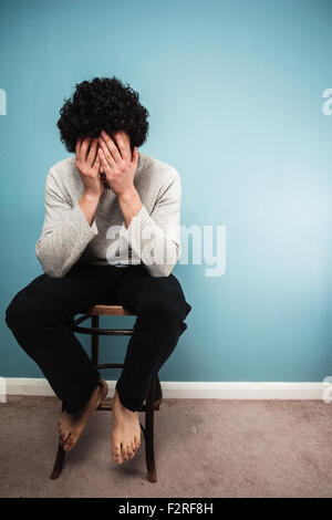 Un jeune homme triste est assis sur une chaise haute par un mur bleu Banque D'Images
