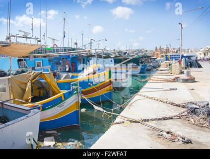 En bois peintes de couleurs vives les petits bateaux amarrés le long du quai sur une journée ensoleillée avec Chursh Paroisse de Marsaxlokk dans la distance. Banque D'Images