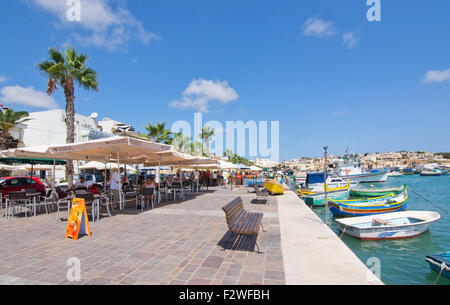 Le restaurant Pier Marsaxlokk avec parasols, bateaux colorés et des signes sur une journée ensoleillée, à Malte. Banque D'Images