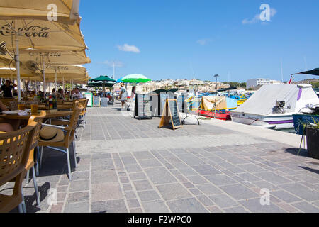 Le restaurant Pier Marsaxlokk avec parasols, bateaux colorés et des signes sur une journée ensoleillée, à Malte. Banque D'Images