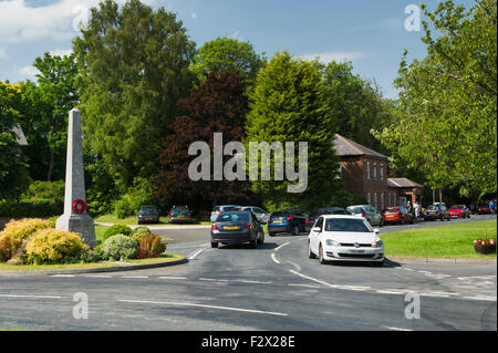 Village le trafic passant un obélisque en pierre (WW1 War Memorial avec couronne de pavot) sur un petit rond-point dans la région ensoleillée Poppleton, près de York, Angleterre, Royaume-Uni. Banque D'Images