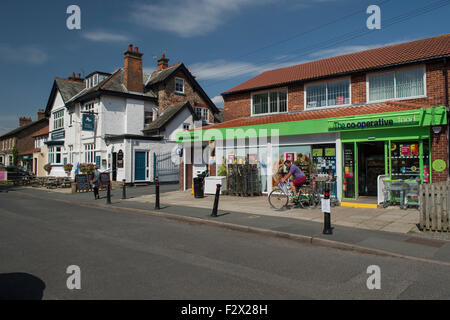 Scène de village - homme vélo loin de Co-operative Food convenience store & dame marchant le long de la route par pub - East Poppleton, York, Angleterre, Royaume-Uni. Banque D'Images