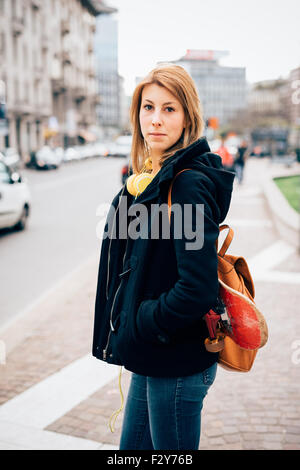 La figure du genou de belle jeune femme blonde sportive hipster posant dans la rue, les mains en poche, à la recherche, à huis clos avec des écouteurs autour de sa poitrine et de skate - musique en beauté, concept Banque D'Images