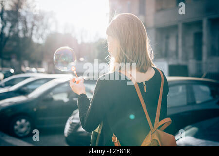 Voir la demi-longueur du dos de la belle jeune femme blonde sportive hipster en ville à jouer avec du savon bulle - l'enfance, le bonheur, l'insouciance concept - rétro-éclairage Banque D'Images
