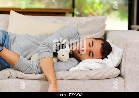 Hispanic male wearing jeans pull bleu et blanc couché sur canapé avec animal en peluche entre les bras de dormir Banque D'Images