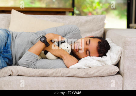 Hispanic male wearing jeans pull bleu et blanc couché sur canapé avec animal en peluche entre les bras de dormir Banque D'Images