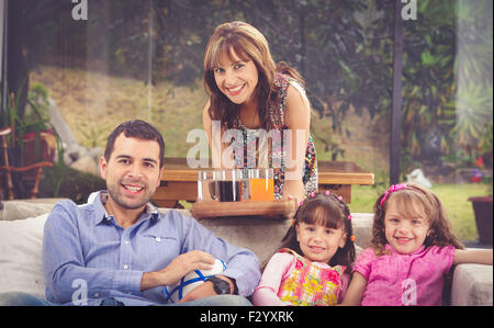 Hispanic father assis dans un canapé avec deux filles et la mère se penche au-dessus de l'arrière plateau de service de boissons, jardin contexte Banque D'Images