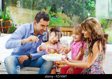 Les parents hispaniques avec deux filles de manger d'un bac de croustilles assis dans un canapé, tandis que de sourire et de profiter de chaque autre société Banque D'Images