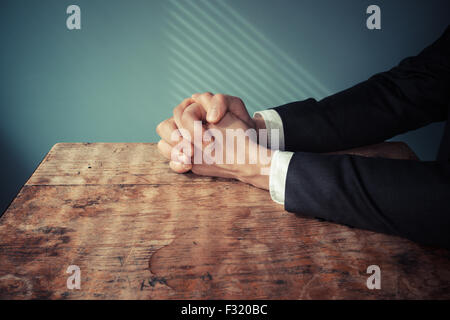 Man in suit avec les mains jointes dans la prière à un bureau avec la lumière pénétrant par les blinds Banque D'Images