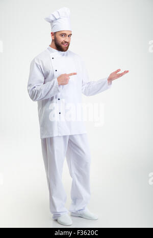 Full Length portrait of a happy chef cook holding copyspace sur le palm et doigt sur elle isolé sur fond blanc Banque D'Images