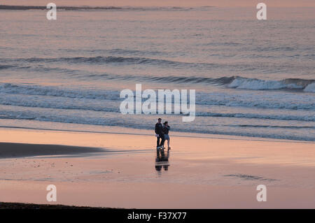 Vue de roman jeune couple marche main dans la main par la mer, au calme, sur une plage de sable déserte au coucher du soleil - Scarborough, Yorkshire Coast, England, UK. Banque D'Images