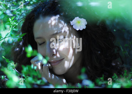 Cute girl entre beaucoup de feuilles vertes avec petite fleur rose tendre dans ses cheveux bouclés Banque D'Images