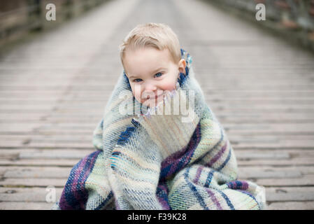 Portrait d'un garçon enveloppé dans une couverture, assis sur un pont en bois