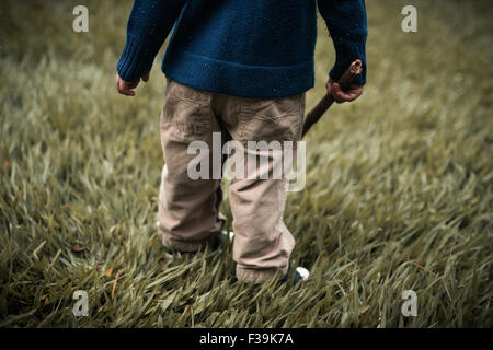 La section d'un enfant en bas standing in field tenant un bâton en bois Banque D'Images