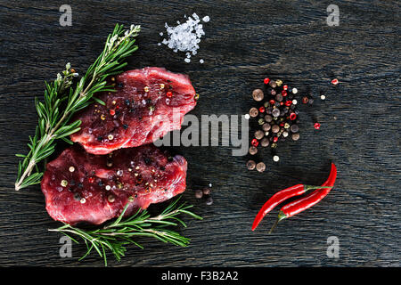 Deux steaks de boeuf garnie avec un couple de brindilles de romarin, nain des piments, sel de mer et poivre en grains sur zone sombre Banque D'Images