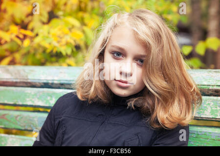 Belle blonde caucasienne teenage girl in black Jacket assis sur le vieux banc vert in autumn park Banque D'Images