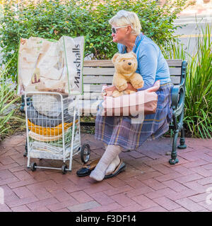 Une femme sans-abri portant des lunettes blanches et la tenue d'un grand ours en peluche animal en peluche est assis sur un banc public. Banque D'Images