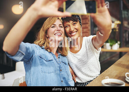 Smiling attractive girlfriends prenant un ensemble selfies, assis dans une cafétéria savourer une tasse de café qui pose pour leur mob Banque D'Images