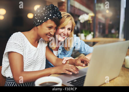 Deux jeunes femmes multiraciales amis en surfant l'internet ensemble sur un ordinateur portable comme ils sont assis dans une cafétéria prendre une tasse de coff Banque D'Images