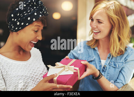 Jolie jeune femme blonde donnant un beau young African American friend un cadeau surprise cadeau avec du papier rouge, un arc d'un Banque D'Images