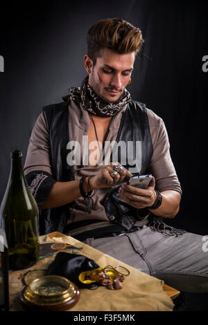 Homme élégant habillé en pirate, avec téléphone cellulaire portant veste en cuir et écharpe assis en chaise à côté de Table, avec vide Bott Banque D'Images