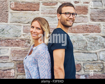 Happy Couple Standing back to back and Smiling at the Camera Against Brick Wall Background Banque D'Images