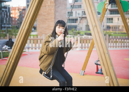 Jeune belle asiatique brune long cheveux droits hipster femme jouant dans un terrain de jeux assis sur une balançoire, à la caméra en souriant, - l'enfance, de la jeunesse, de l'insouciance concept Banque D'Images