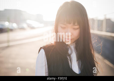 Portrait of young belle asiatique brune long cheveux droits hipster femme dans la ville, à la recherche vers le bas, pensive pendant le coucher du soleil dans la pensée réfléchie - rétroéclairage, avenir, concept graves Banque D'Images