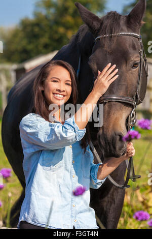 Belle jeune femme asiatique ou eurasienne girl wearing denim shirt, souriant et menant son cheval en soleil Banque D'Images